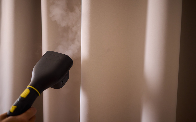 A hand-held steamer being used to sanitize and remove wrinkles from a set of thick, neutral-colored curtains.