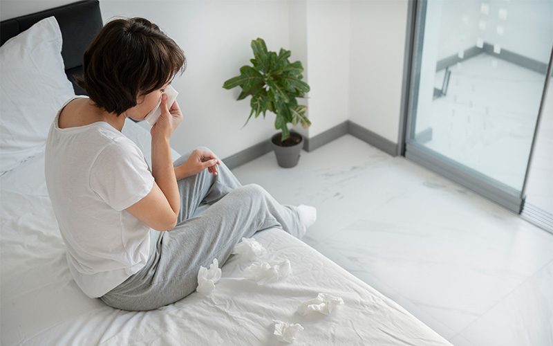 Person in white t-shirt and sweatpants holding tissue, surrounded by used tissues in bright room.