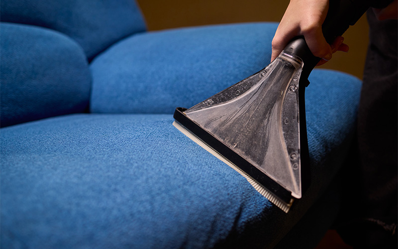 Handheld extractor cleans blue sofa cushion with transparent nozzle tool.