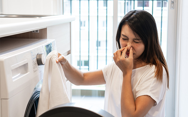 Woman smelling musty laundry from a washing machine at home.