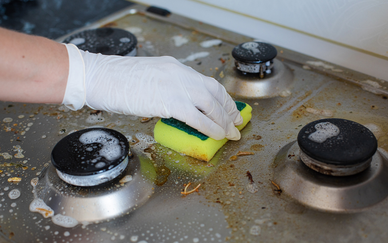 Hands wearing gloves cleaning a greasy gas stove burner